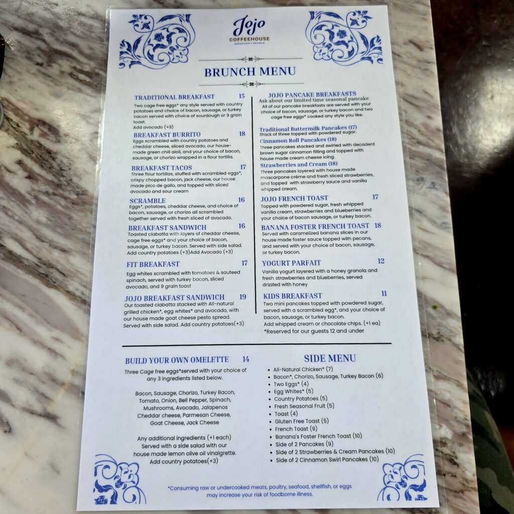 A photo of the JoJo Coffeehouse brunch menu placed on a marble tabletop, showing breakfast items such as pancakes, breakfast burritos, tacos, omelettes, and sides.