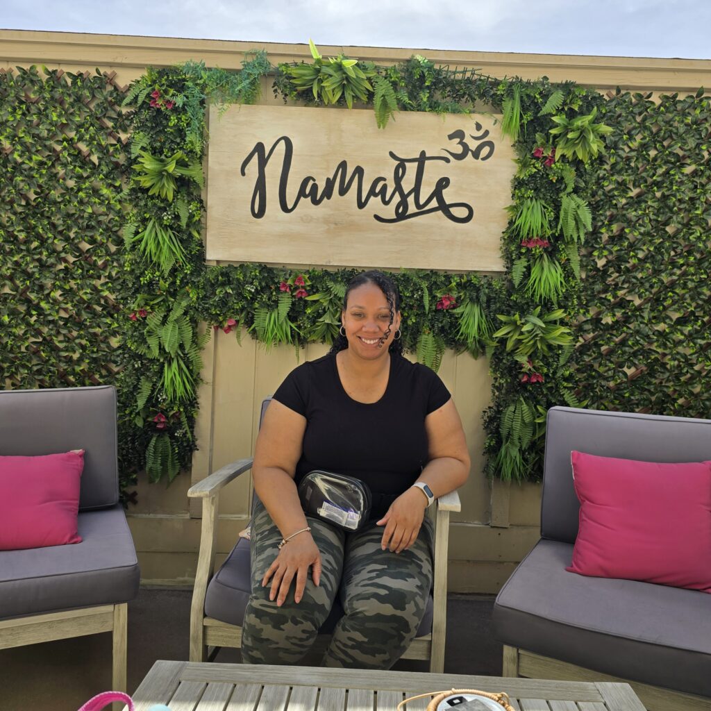 Calesse ‘Curly’ Haynes smiling while seated on an outdoor patio, in front of a lush green plant wall with a wooden sign that reads ‘Namaste.