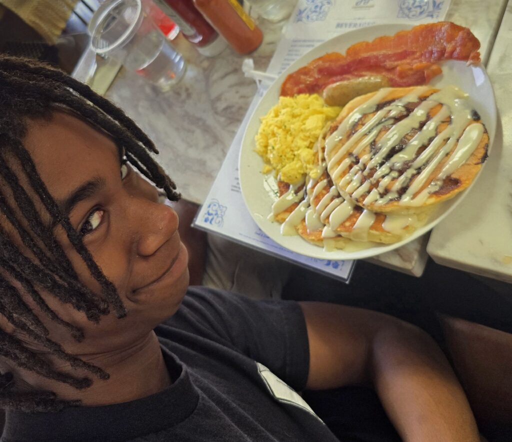 A smiling teen seated at a restaurant table with a plate of cinnamon roll pancakes topped with cream cheese icing, served with scrambled eggs, bacon, and sausage.