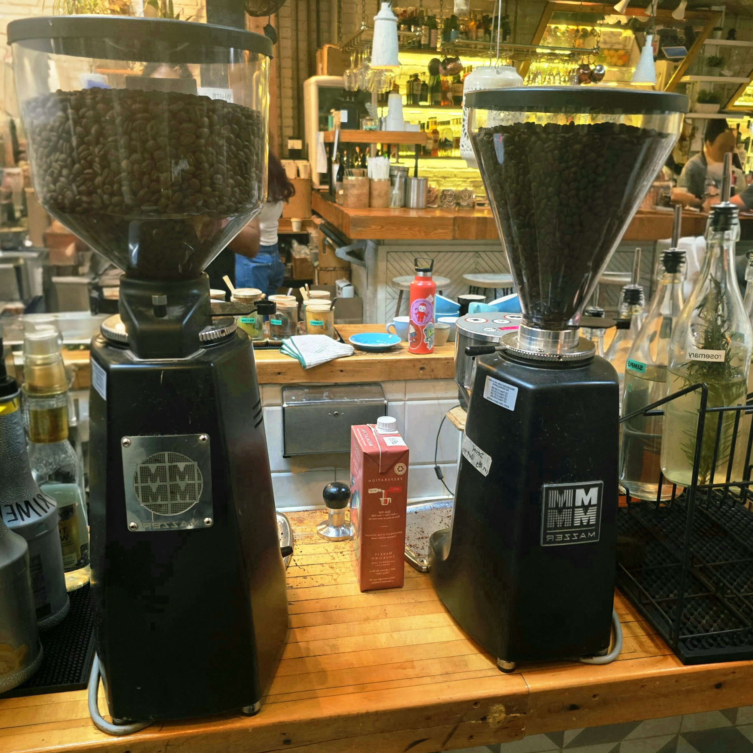Two large commercial coffee grinders filled with whole coffee beans on a wooden café counter, surrounded by syrups, bottles, and barista tools in a cozy, bustling coffee bar.