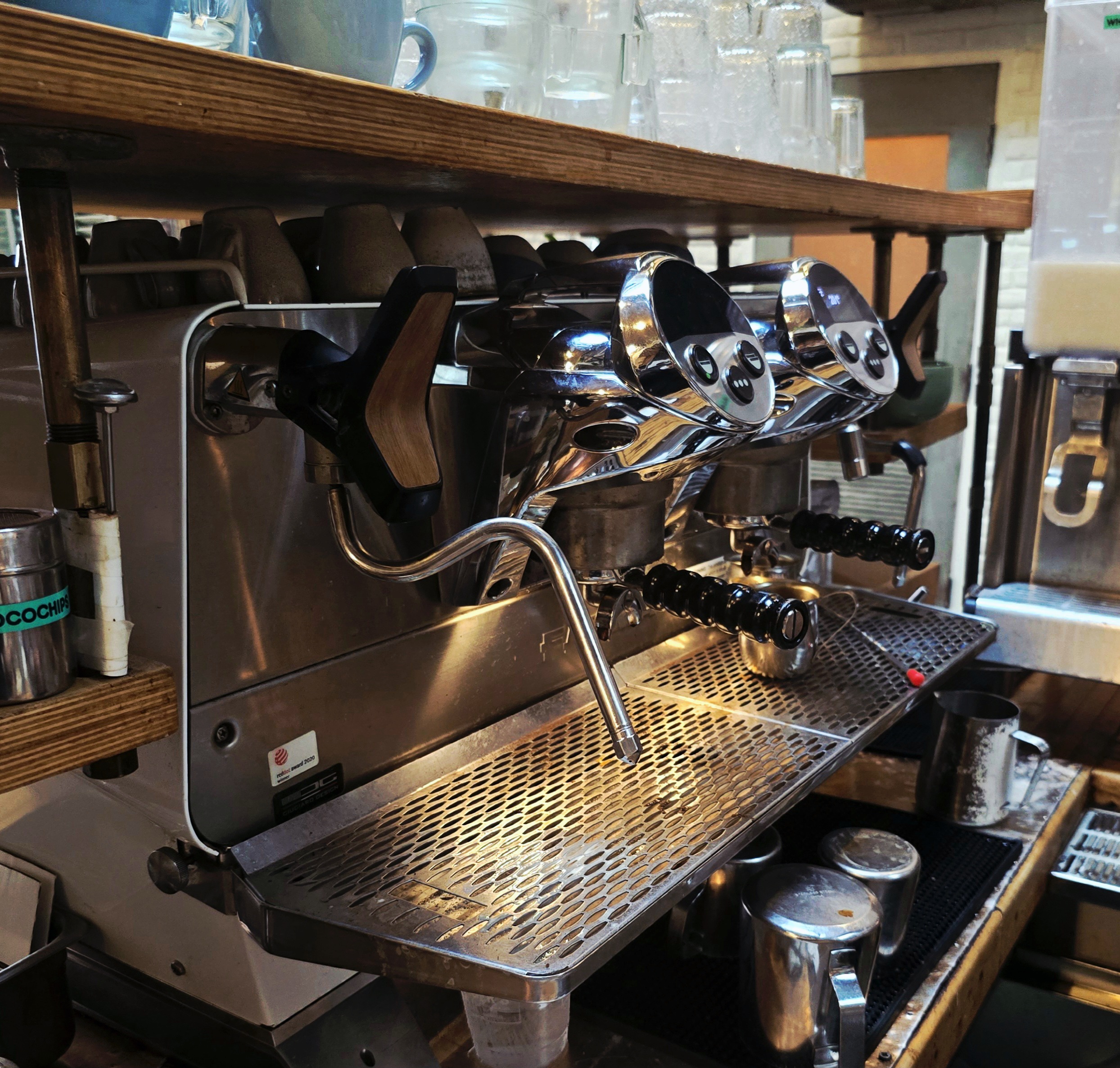 A close-up view of a stainless-steel commercial espresso machine with dual group heads, steaming wand, and metal pitchers arranged on the counter beneath warm café lighting.