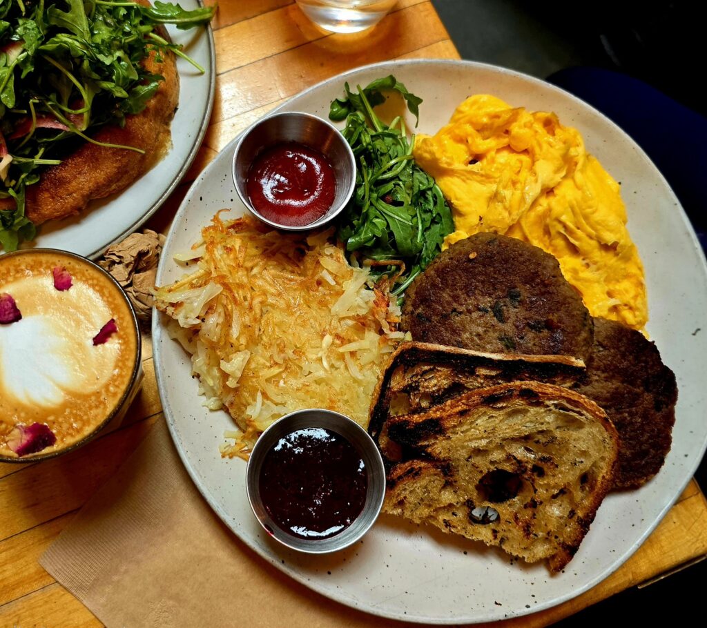 Vegetarian country-style breakfast with fluffy scrambled eggs, crispy hash browns, plant-based sausage patties, toasted rustic bread, fresh greens, and jam on a wooden table.
