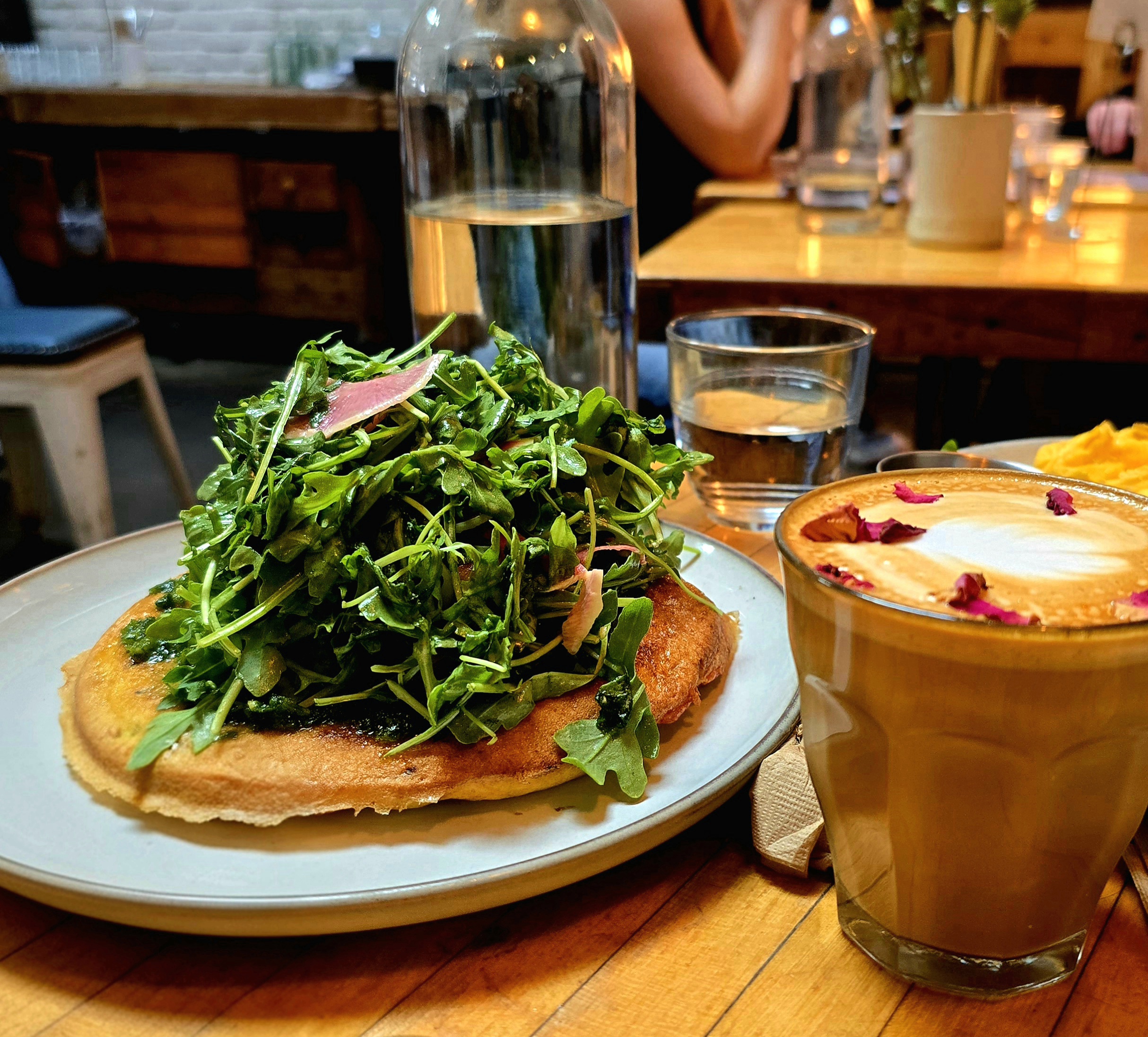 A plate with a golden frittata topped with a generous pile of fresh arugula and pickled onions, served alongside a rose-topped latte on a wooden café table with water glasses and soft ambient lighting in the background.
