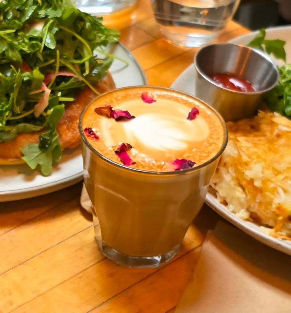 A rose cardamom latte topped with dried rose petals on a wooden café table, surrounded by brunch dishes including hash browns, greens, and a frittata plate.