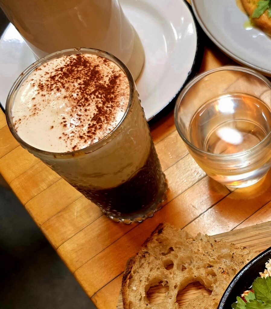 A tall glass of salted cream coffee topped with a thick, frothy layer dusted in cocoa powder, sitting on a wooden café table next to a glass of water and a slice of toasted sourdough.