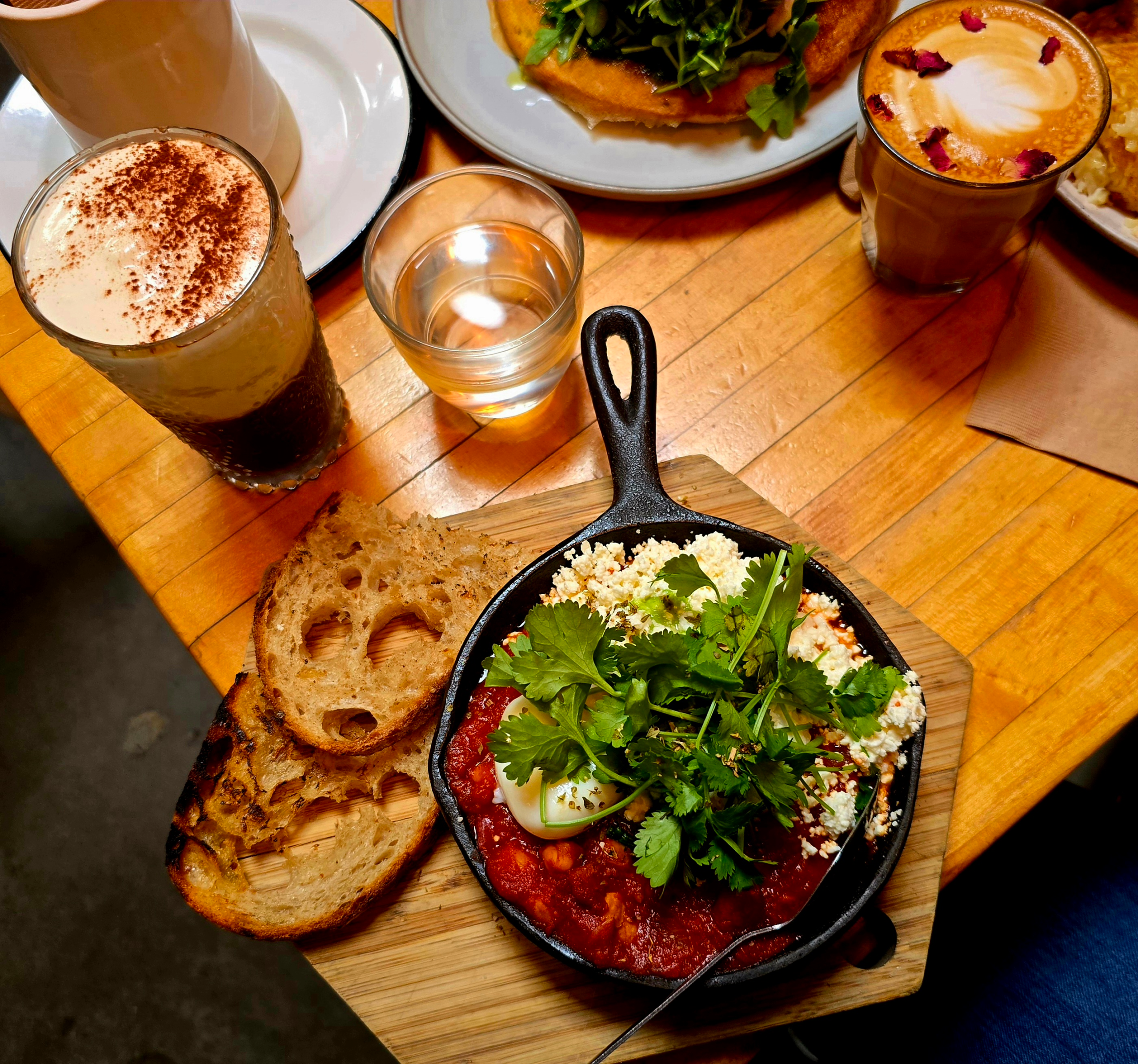 A skillet of shakshuka topped with poached eggs, cilantro, and crumbled cheese, served with slices of toasted sourdough on a wooden board, surrounded by lattes and other brunch dishes on a wooden café table.