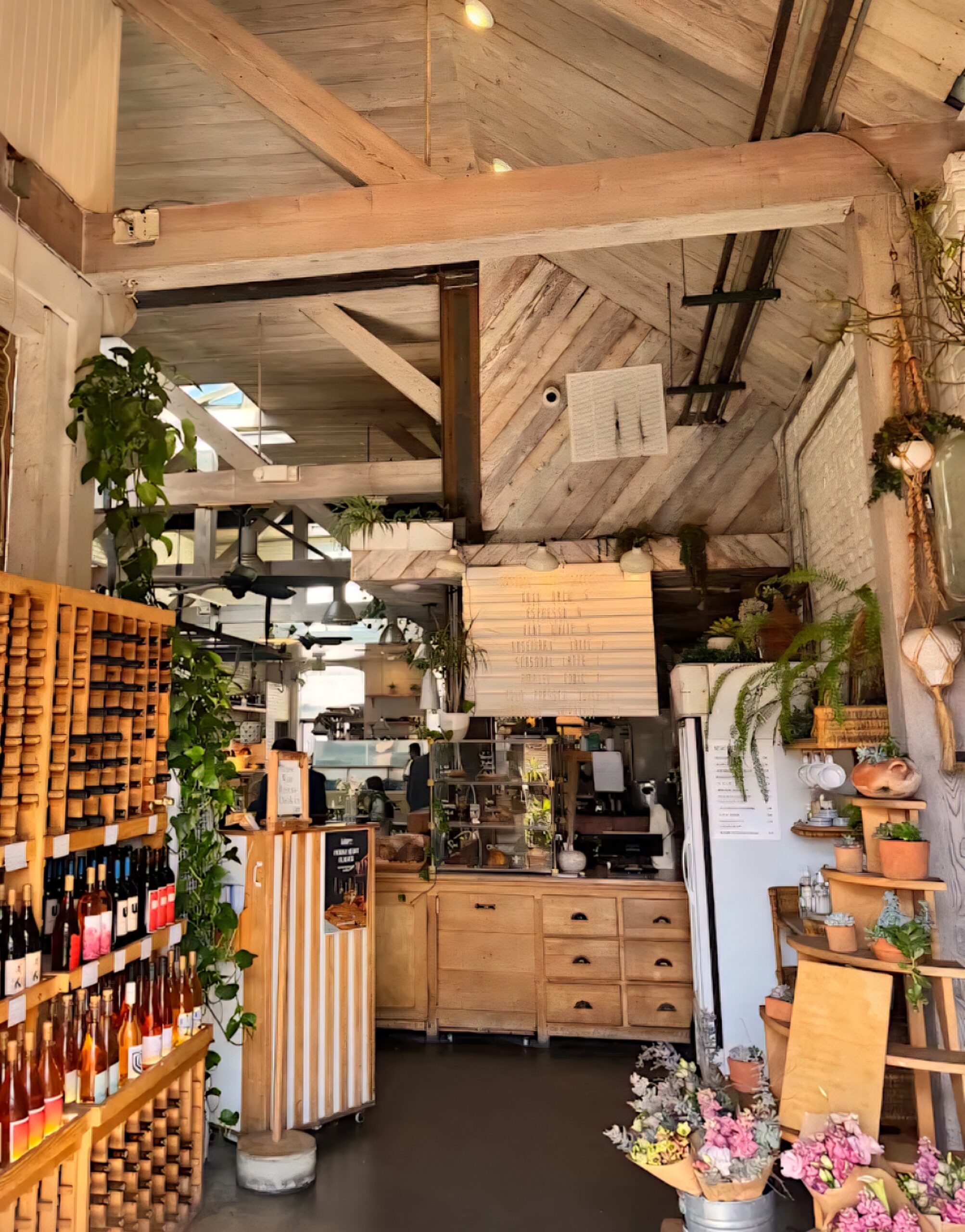 A bright, rustic café interior at The Butcher’s Daughter featuring warm wood tones, hanging plants, shelves of wine bottles, a floral display, and a cozy counter area with natural light streaming through the ceiling beams.