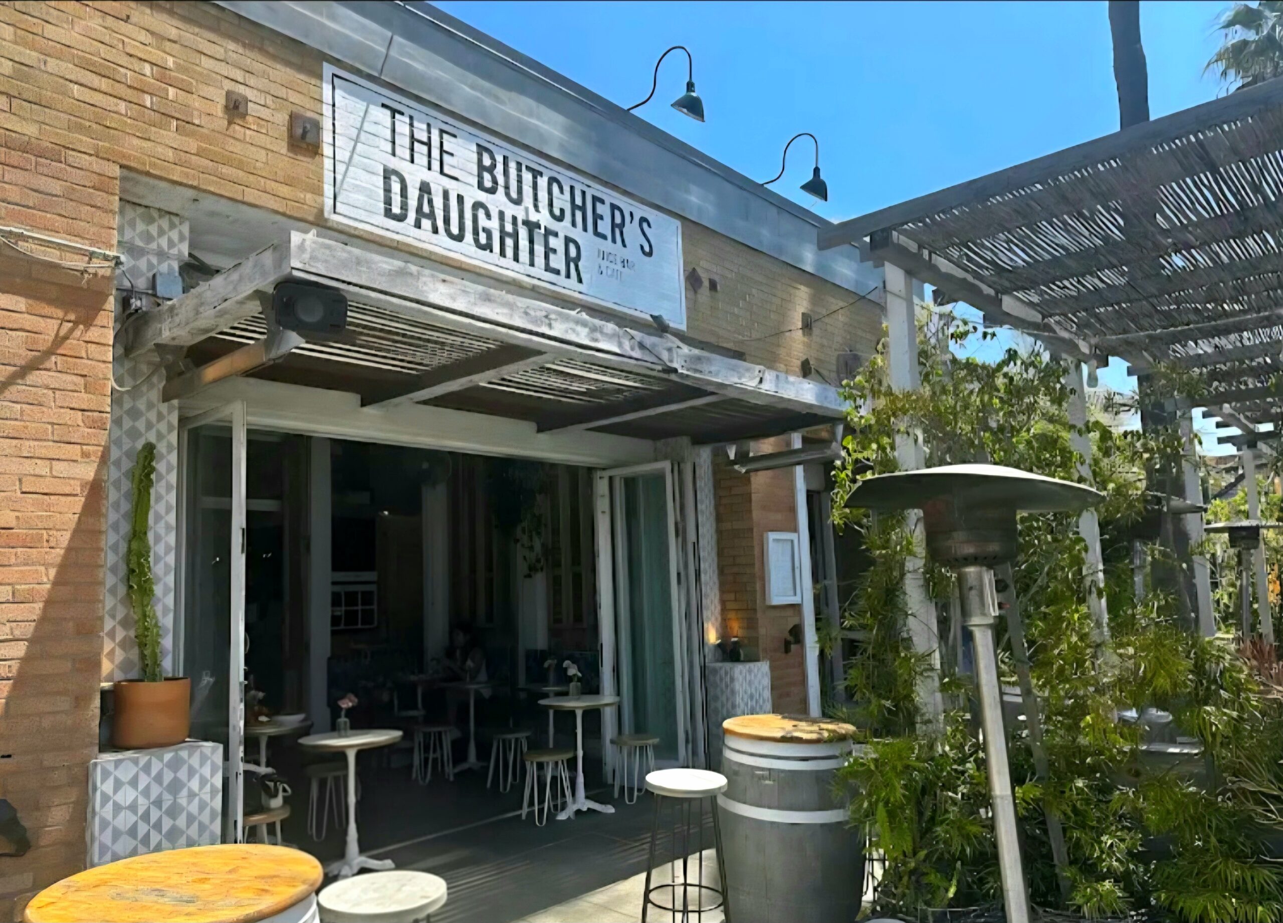 Exterior of The Butcher’s Daughter restaurant with open-air seating, rustic wood accents, and greenery under a bright sunny sky in Los Angeles.