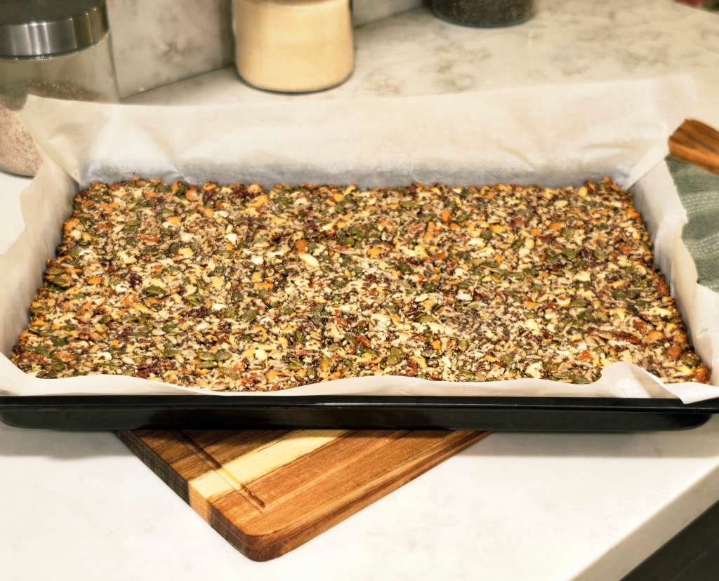 A full sheet of baked seeded brittle in a parchment-lined baking pan, cooling on a wooden cutting board on a kitchen counter.