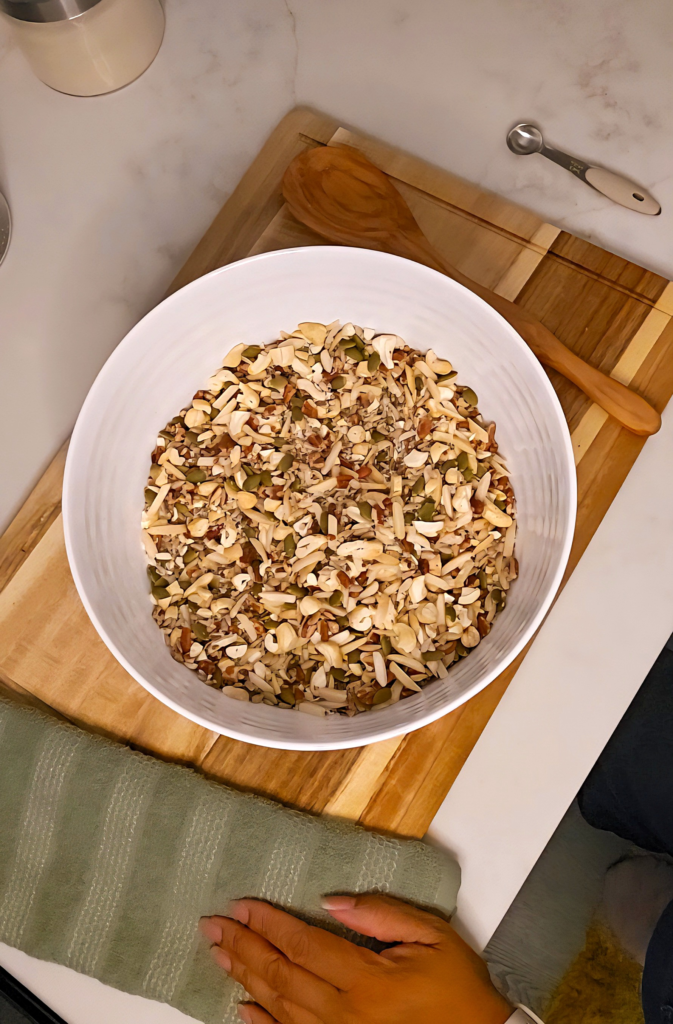 A large white mixing bowl filled with chopped nuts and seeds on a wooden cutting board, with a wooden spoon, a measuring spoon, and a green kitchen towel with a hand resting on it nearby.