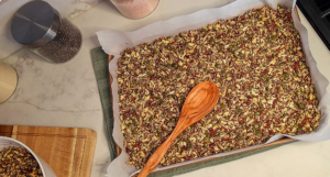 A baking sheet lined with parchment and filled with an even layer of homemade seeded brittle mixture, topped with pumpkin seeds, pecans, and chia, with a wooden spoon resting on top on a marble countertop.