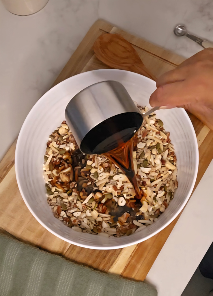 A hand pours maple syrup from a metal measuring cup into a bowl of mixed nuts and seeds on a wooden cutting board.