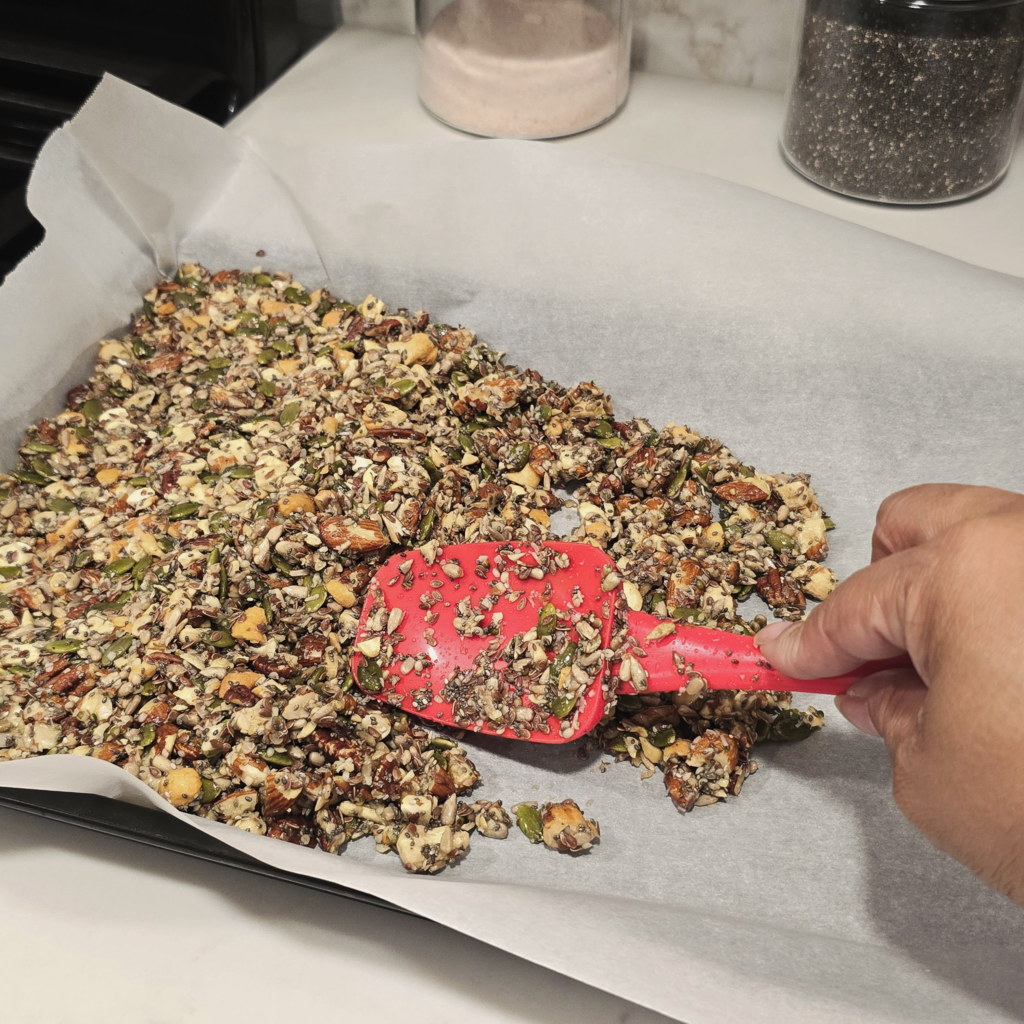 A hand uses a red spatula to spread maple-coated nuts and seeds evenly across a parchment-lined baking sheet.