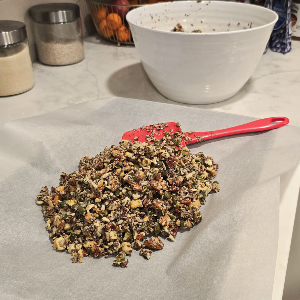 A mound of maple-coated nuts and seeds sits on a parchment-lined baking sheet with a red spatula beside it, while a mixing bowl rests in the background.