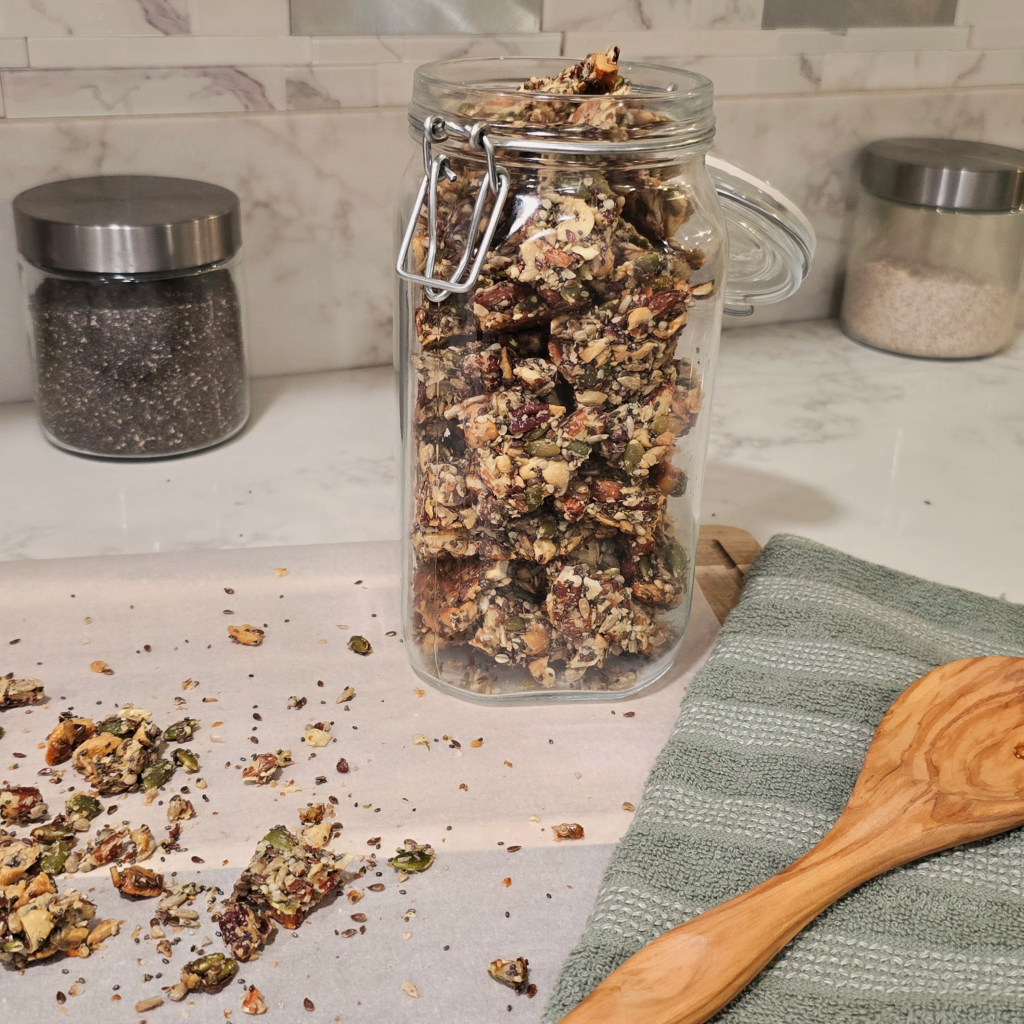 A tall glass jar filled with pieces of homemade seeded brittle stored on parchment paper, with a green kitchen towel and wooden spoon nearby on a marble countertop.