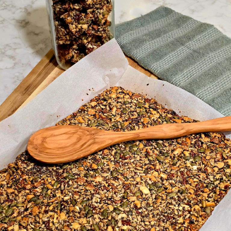 A freshly baked sheet of homemade seeded brittle resting on parchment paper with a wooden spoon on top, set on a marble countertop with a jar of brittle pieces and a folded green kitchen towel nearby.