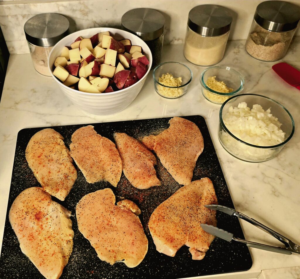 Seasoned raw chicken breasts arranged on a cutting board alongside chopped onions, garlic, and prepared ingredients.